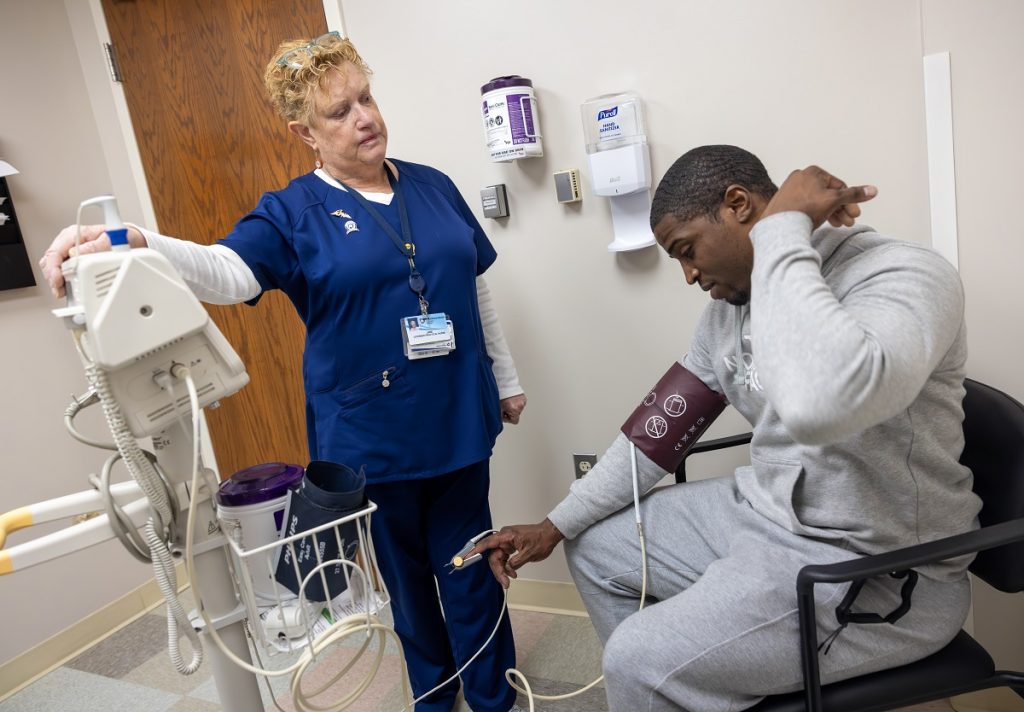 Jane Pizarro, a licensed practical nurse at Penn State Health St. Joseph Downtown Campus, listens to patient Kelvin Bernard Tyler Jr. She is wearing scrubs, a long-sleeved shirt, lanyard and name badge and has glasses on her head. He is wearing sweatpants and a sweatshirt and is gesturing toward his neck. He has a blood pressure cuff on his arm and a pulse monitor on his finger.