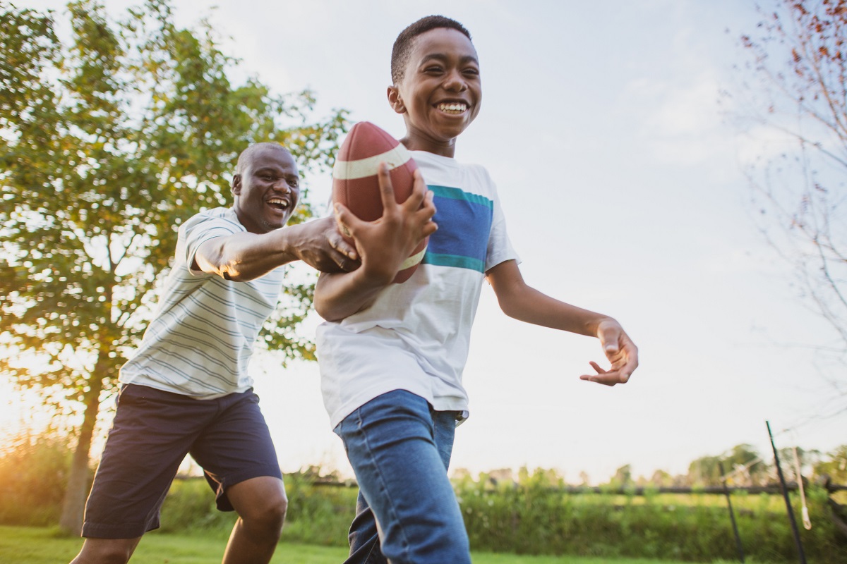 A boy and his dad laugh together while playing football on a lawn in the summertime.