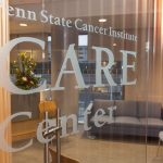 Double glass doors with a decal that says, “Penn State Cancer Institute CARE Center.” Through the doors there is a chair, a sofa and a bouquet of flowers on a counter.