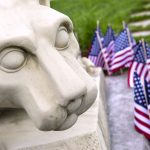 Close-up of the Nittany Lion statue’s face with small flags on the right. Paving stones and lawn are also on the right.