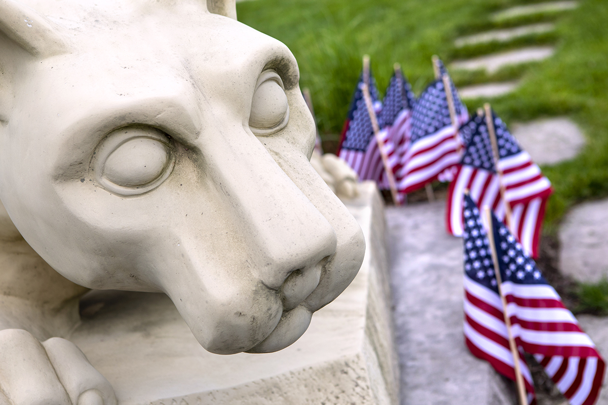 Close-up of the Nittany Lion statue’s face with small flags on the right. Paving stones and lawn are also on the right.