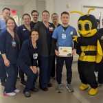 A male patient care associate wearing hospital scrubs holds a certificate honoring him for great patient care. He is surrounded by ten male and female hospital teammates. A person dressed in a bee costume points to the honoree.