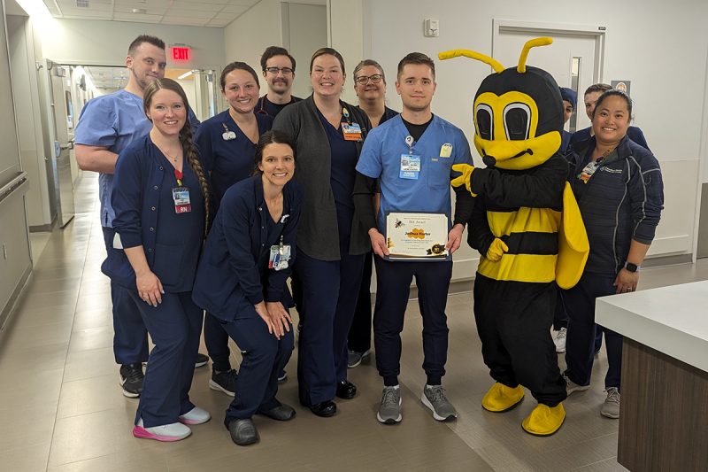 A male patient care associate wearing hospital scrubs holds a certificate honoring him for great patient care. He is surrounded by ten male and female hospital teammates. A person dressed in a bee costume points to the honoree.