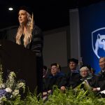 Kristen Manto, in commencement cap and gown, speaks behind a lectern with the Penn State shield while faculty sit in a row of chairs behind.