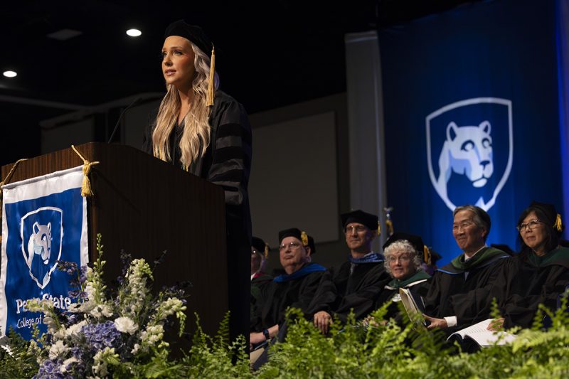 Kristen Manto, in commencement cap and gown, speaks behind a lectern with the Penn State shield while faculty sit in a row of chairs behind.