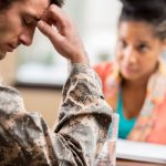 Side view of gentleman in military camo holding his head. Concerned woman in background looking at him with notebook in front of her.