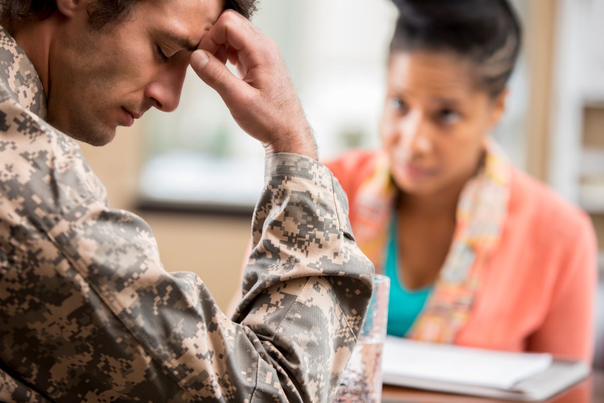 Side view of gentleman in military camo holding his head. Concerned woman in background looking at him with notebook in front of her.