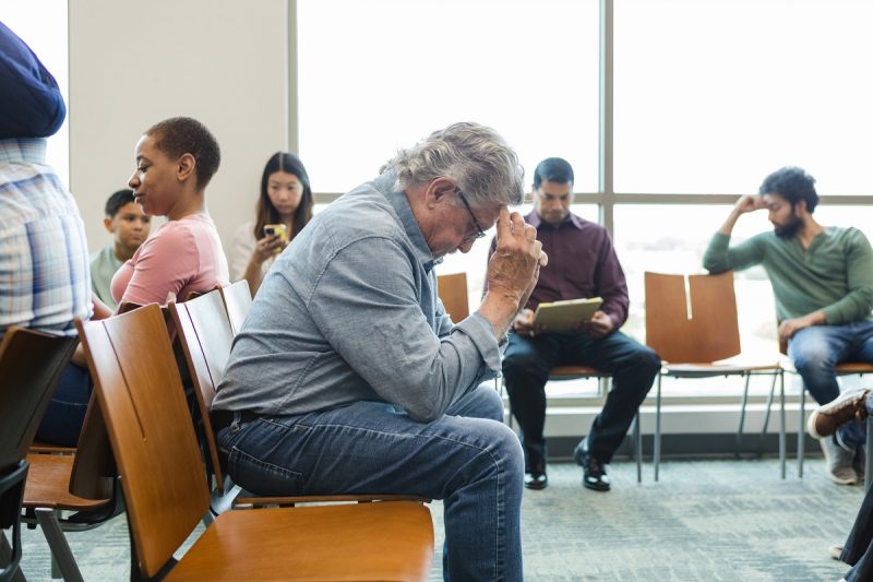 An older adult man sits anxiously in a hospital waiting room with his hands against his forehead.