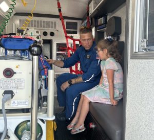A flight paramedic and a young girl sit in the back of an ambulance, as the paramedic explains to her the various pieces of equipment that surround them.