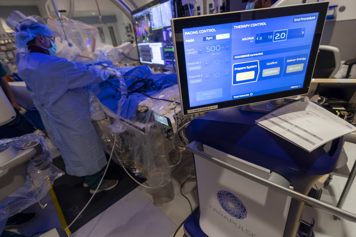 A physician wearing a cap, mask and gown performs a procedure on a patient, referencing a nearby monitor. Another monitor is located closer to the camera.