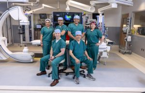 Six physicians in scrubs – four standing, two sitting – pose for a photo in a medical procedure room.