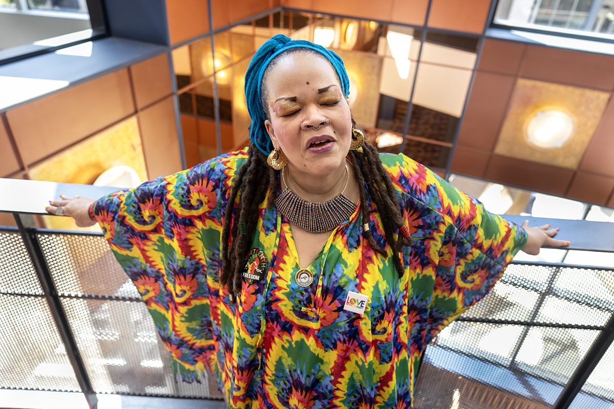 A woman wearing a tie-dyed shirt, dread locks, and a bandana on her head sings with eyes closed, leaning back on a railing with arms stretched out.