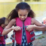 Young girls put on life jackets while standing along the side of a lake.