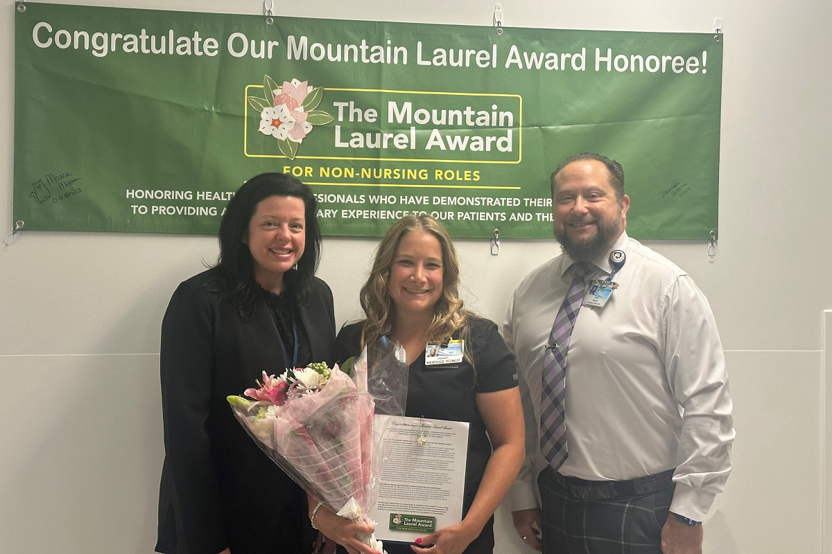 Two ladies and a gentleman stand side-by-side, smiling. The lady in the middle holds a certificate and flowers. On the wall above and behind them is a banner that says, “Congratulate Our Mountain Laurel Award Honoree! The Mountain Laurel Award for non-nursing roles.”