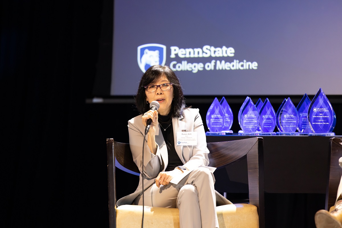 A woman sits on a stage at an awards program, holding a microphone and speaking to an audience