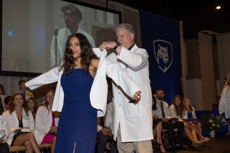 A man wearing a white doctor's coat cloaks a young women in a shorter doctor's coat, marking the beginning of medical school