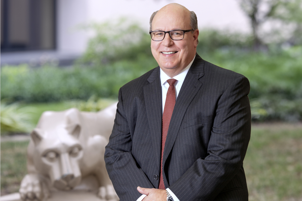 Steve Massini poses for a photo wearing a suit and seated near a Nittany Lion statue.