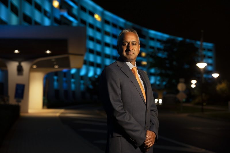 A man poses for a portrait in front of a building. The building is illuminated in blue lights.