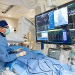 A physician in an operating room wearing scrubs and glasses stands over a patient and looks at a computer monitor.