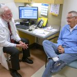 Two men sit next to a desk, while looking at each other with their hands clasped together. The man on the left wears glasses and a lanyard around his neck. A stethoscope is in the pocket of his jacket. The man on the right sits with one leg crossed over the other. A computer, keyboard, phone, papers, folders and market sit on the desk behind them.