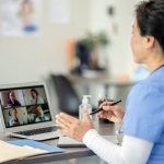 A female clinician dressed in a medical smock sits with her back to the camera, a computer screen on the desk to her left in front of her. On the computer screen are the images of four other people involved in the educational session including those in a clinical setting and in an office.