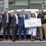 A group of people stand in front of a large SUV, holding a large check and smiling