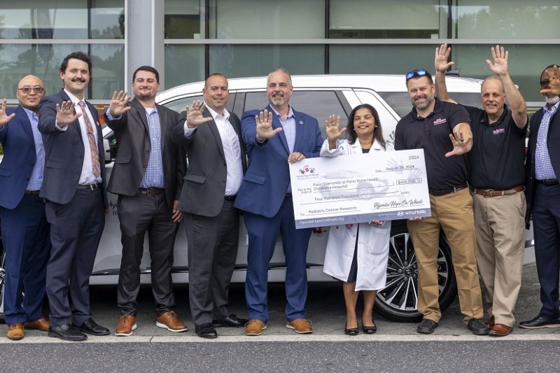 A group of people stand in front of a large SUV, holding a large check and smiling