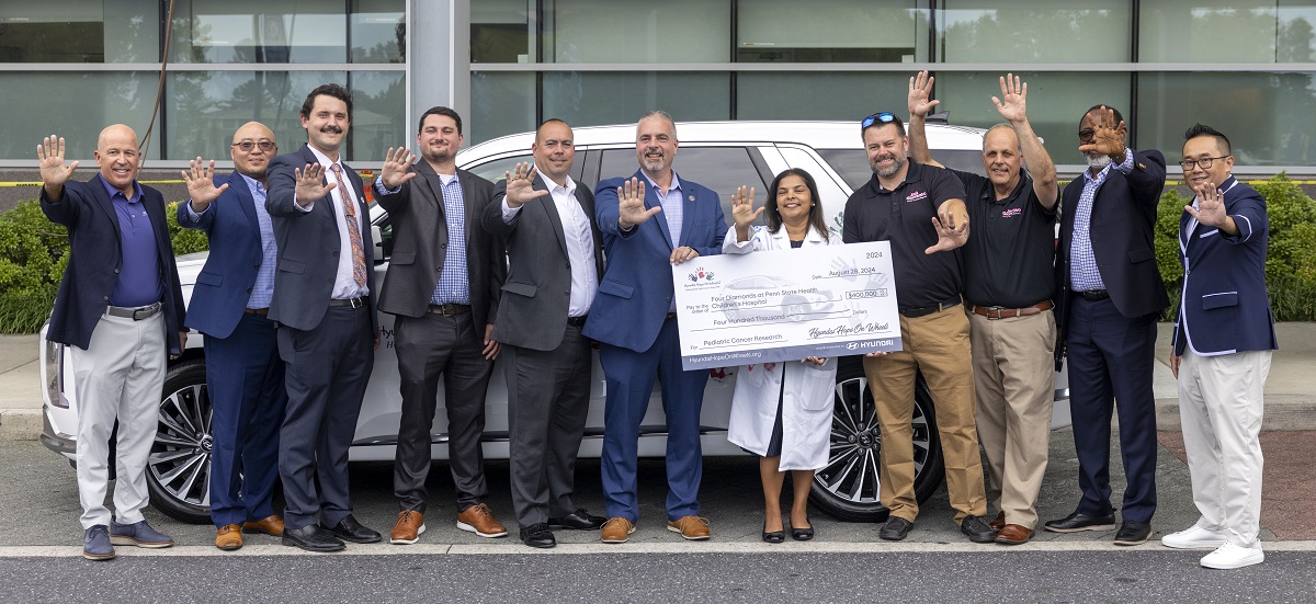 A group of people stand in front of a large SUV, holding a large check and smiling