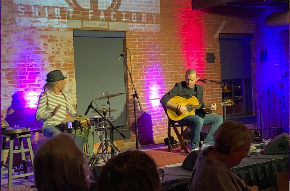 Musicians, seated, play on a stage; an acoustic guitarist is on the right and a drummer on the left