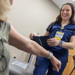 a certified medical assistant at Penn State Medical Group – assists a patient by taking their blood pressure. She is wearing blue scrubs and a name badge.