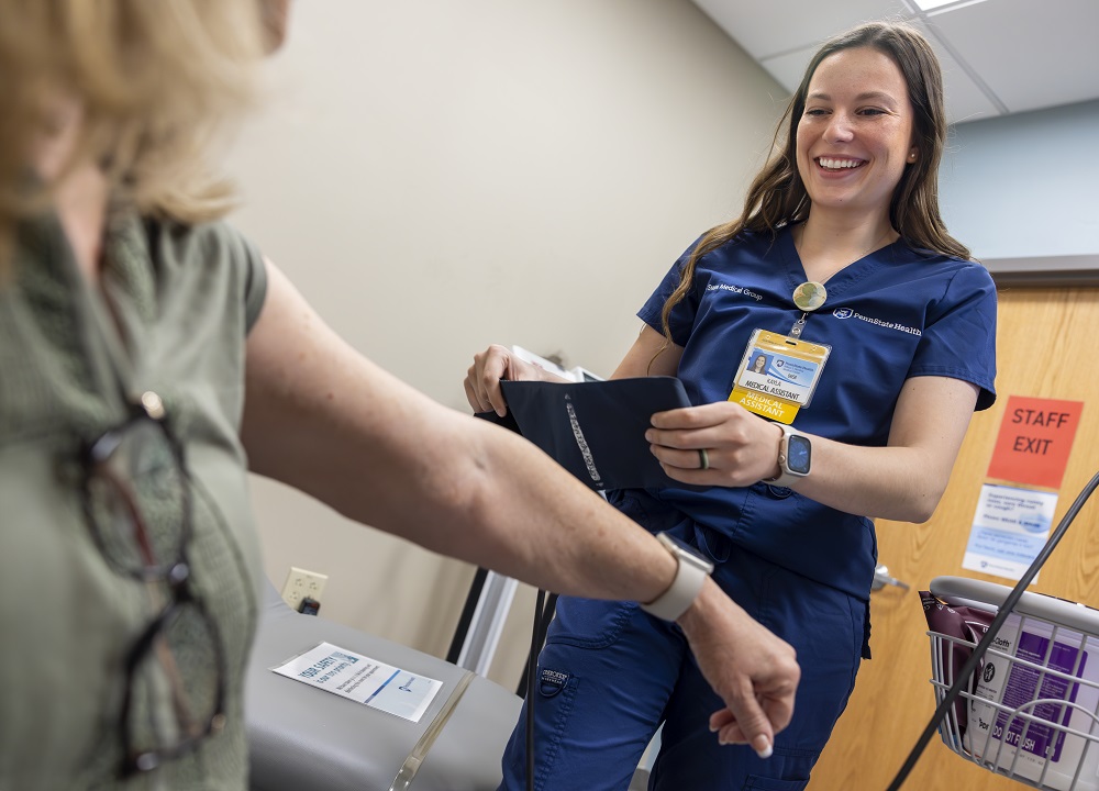 a certified medical assistant at Penn State Medical Group – assists a patient by taking their blood pressure. She is wearing blue scrubs and a name badge.