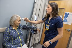 A certified medical assistant at Penn State Health Medical Group – Cocoa, takes a patient's temperature using a forehead thermometer. She is wearing blue scrubs and a name badge. The patient, an older woman, sits calmly in a chair.