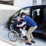 An older man with glasses and a sweater is helped into a wheelchair next to the man’s van, parked in an accessible parking space at a hospital. A valet assistant, dressed in pants and a pullover shirt with a lanyard, is bending over the man, helping him into the wheelchair.