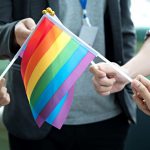 Several individuals, none of whose face can be seen, hold small Pride flags at an outside event.