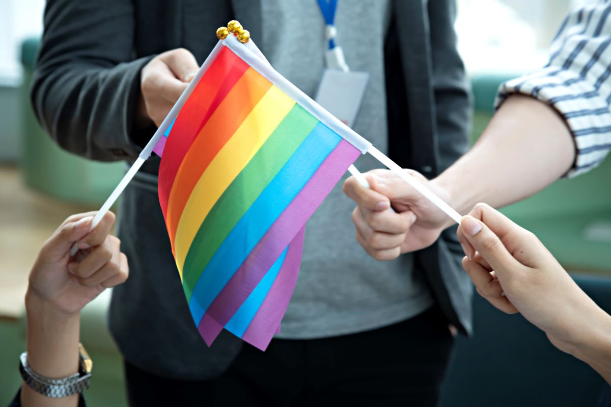Several individuals, none of whose face can be seen, hold small Pride flags at an outside event.