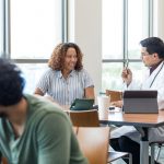 Several people are seated at tables in front of large windows. All have computer-type devices. In the middle table are a woman, who is wearing a striped shirt and has wavy hair, and a man in a medical coat who are discussing the material they are reviewing on screen. The man is holding a pen as if to make a point.