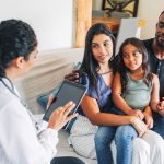 A Hispanic/Latino family sits on a couch listening to a female health care provider, who sits to the left of them and has her back to us. She has a stethoscope around her neck and is reviewing information on a portable computer tablet. She has long curly hair. The family consists of a mother, who also has long hair and is dressed in slacks and a blouse, holding a girl of about seven or eight years of age on her lap. To the right of the woman is a man who has a beard and is wearing jeans and a pullover shirt. He is holding the child’s left hand.