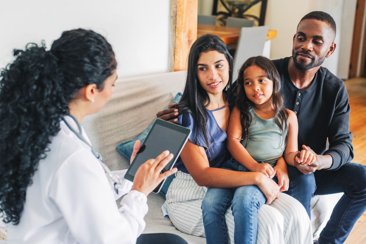 A Hispanic/Latino family sits on a couch listening to a female health care provider, who sits to the left of them and has her back to us. She has a stethoscope around her neck and is reviewing information on a portable computer tablet. She has long curly hair. The family consists of a mother, who also has long hair and is dressed in slacks and a blouse, holding a girl of about seven or eight years of age on her lap. To the right of the woman is a man who has a beard and is wearing jeans and a pullover shirt. He is holding the child’s left hand.