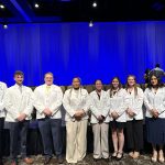 A group of nine college students wearing white medical coats stand together in front of a stage.