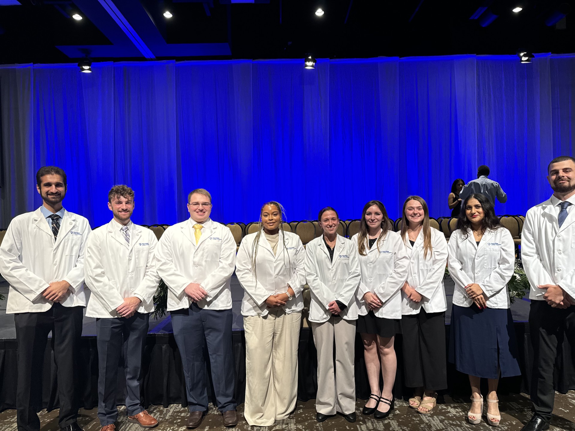 A group of nine college students wearing white medical coats stand together in front of a stage.