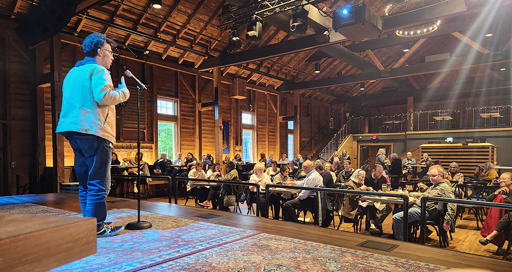 Jason Plotkin speaking behind a microphone on a stage with audience sitting at tables in front