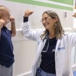 Dr. Sholler, wearing a lab coat and glasses, smiles as she raises both arms to flex her biceps. A boy next to her smiles back and mimics her.