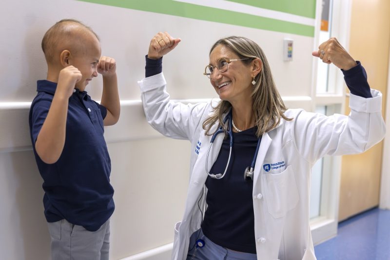 Dr. Sholler, wearing a lab coat and glasses, smiles as she raises both arms to flex her biceps. A boy next to her smiles back and mimics her.