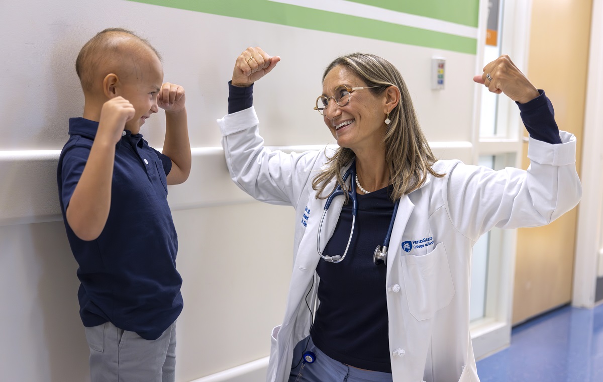 Dr. Sholler, wearing a lab coat and glasses, smiles as she raises both arms to flex her biceps. A boy next to her smiles back and mimics her.