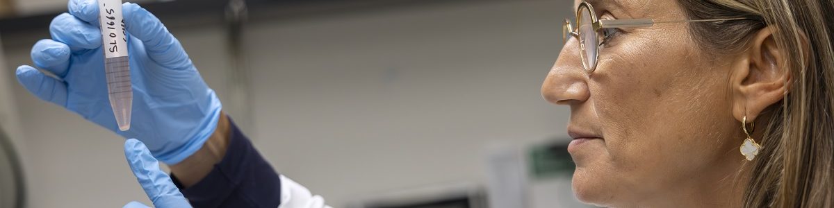 Dr. Sholler holds up a test tube with hands sheathed in rubber gloves. She examines the contents inside.