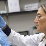 Dr. Sholler holds up a test tube with hands sheathed in rubber gloves. She examines the contents inside.