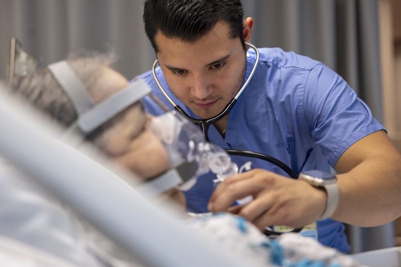 Dr. Raul Vazquez Urrutia, left, listens to the heart of a patient in the Medical Intensive Care Unit of Penn State Health Milton S. Hershey Medical Center. He has a stethoscope in his ears and is wearing scrubs. The patient is lying in a hospital bed and wearing an oxygen mask.