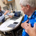 Tyler Thomas, left, gestures as he talks to patient John Jones, right. Thomas is wearing a button-down shirt, tie, glasses and has a stethoscope around his neck. Jones is wearing a Hawaiian shirt and has his hands over his heart.