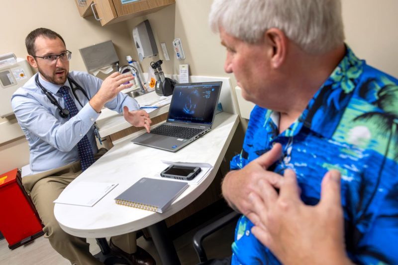 Tyler Thomas, left, gestures as he talks to patient John Jones, right. Thomas is wearing a button-down shirt, tie, glasses and has a stethoscope around his neck. Jones is wearing a Hawaiian shirt and has his hands over his heart.