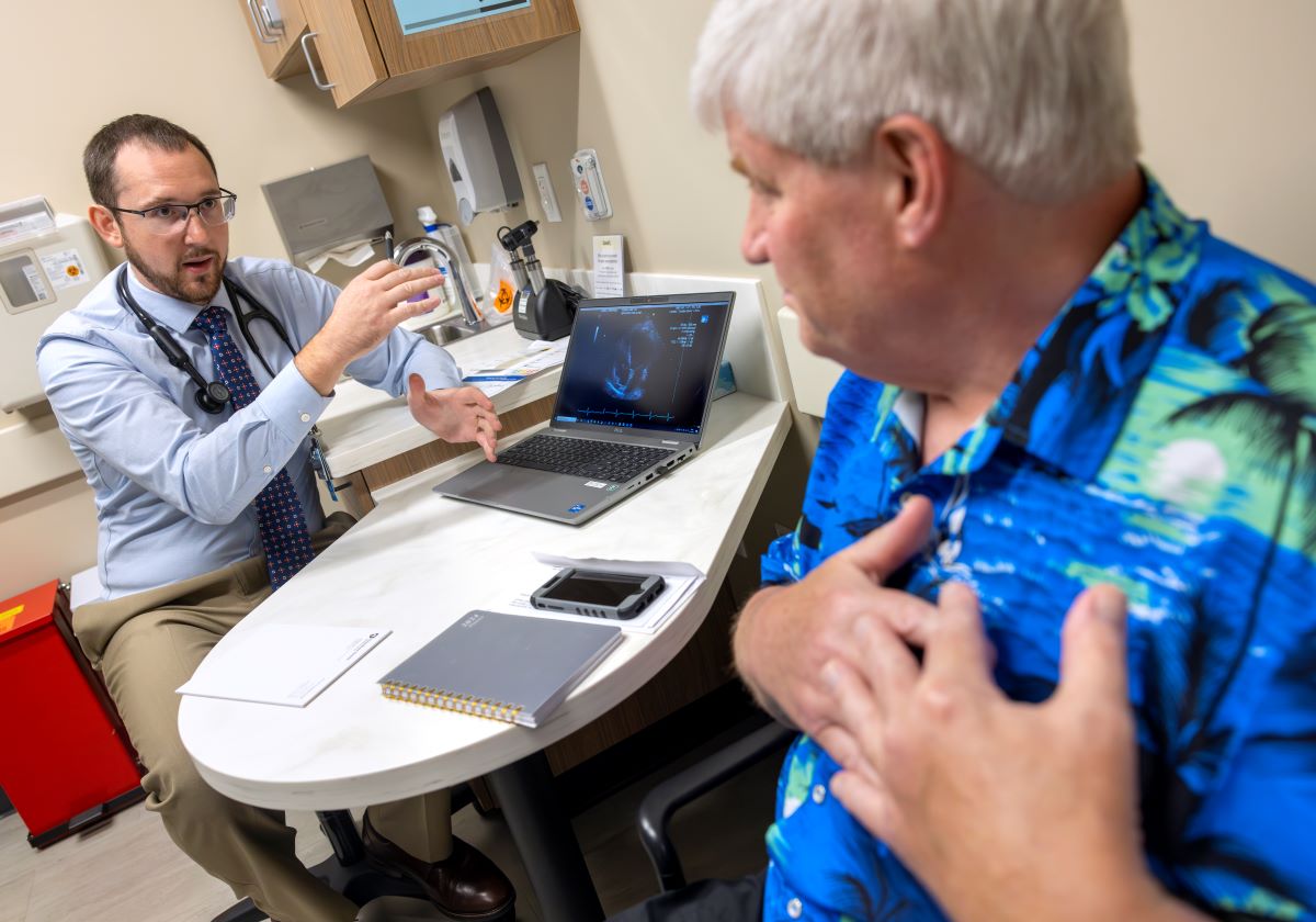 Tyler Thomas, left, gestures as he talks to patient John Jones, right. Thomas is wearing a button-down shirt, tie, glasses and has a stethoscope around his neck. Jones is wearing a Hawaiian shirt and has his hands over his heart.